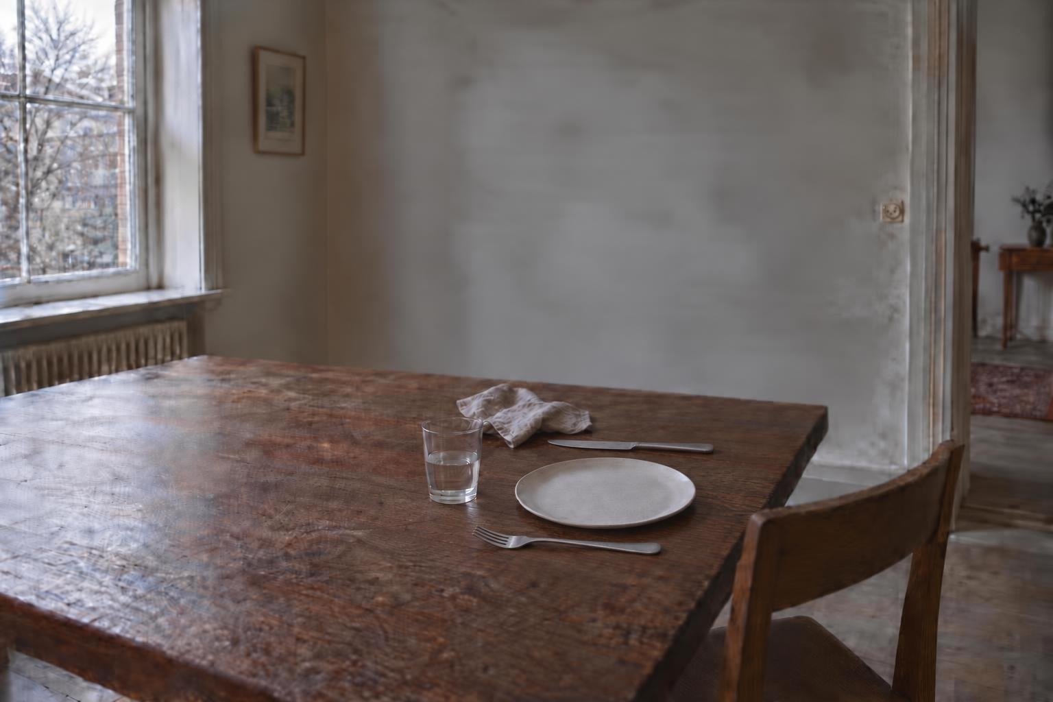 Illustration: a table by a window in grey morning light, set for one, with a glass of water and a chair pulled out.